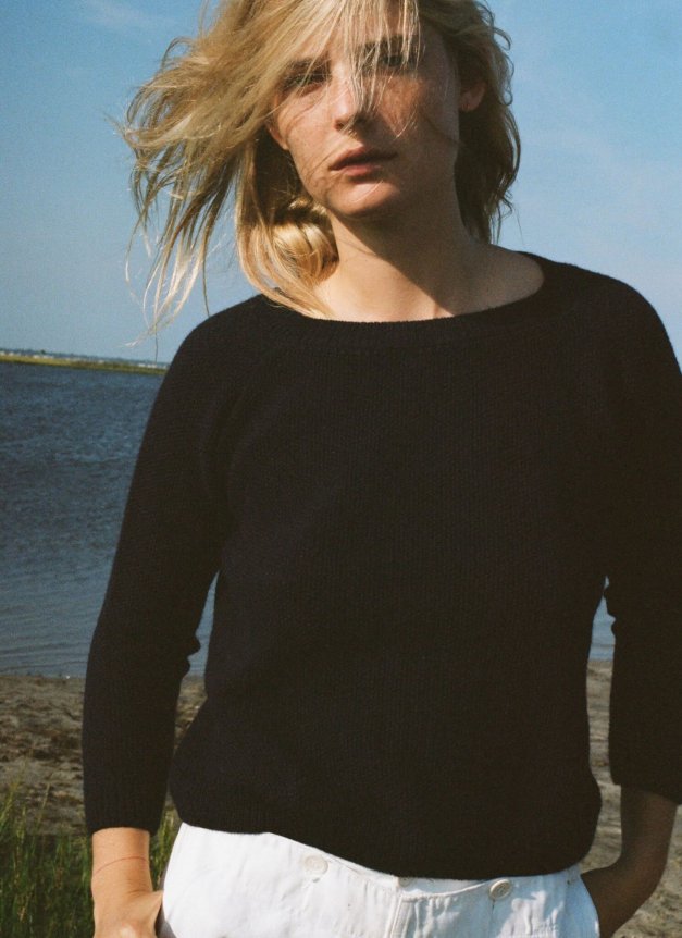 A woman wearing a black fitted, long-sleeve knit sweater and white high-waisted pants stands by a waterfront with windblown hair.