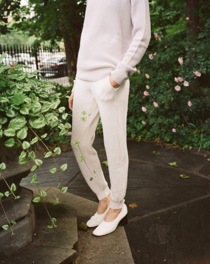 A person wearing an elegant, cream knit sweater and pants ensemble with a relaxed fit, complemented by white block-heeled shoes, set in a lush garden backdrop.
