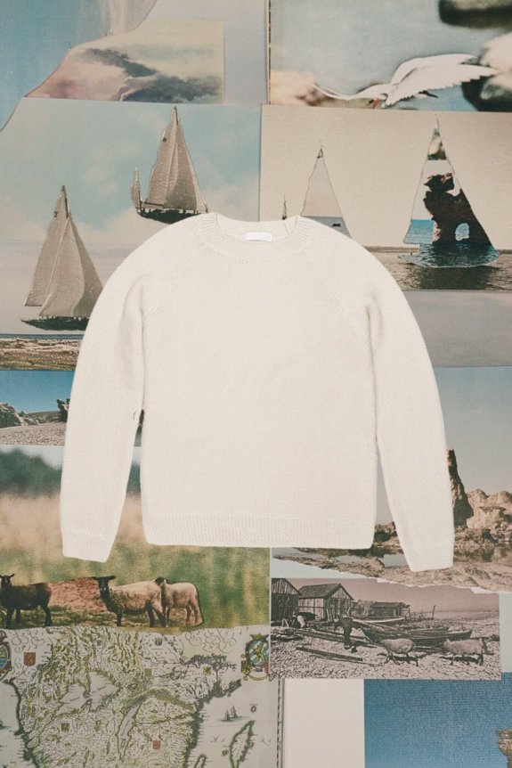 A cream-colored, long-sleeve, crewneck sweater with a textured knit pattern, set against a collage of nautical and pastoral images.
