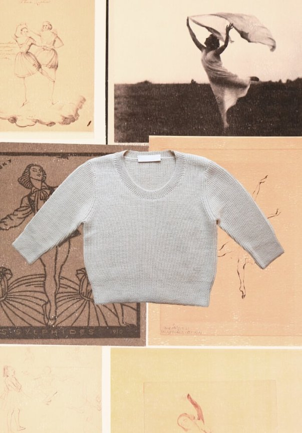 A soft gray knit sweater with a simple, classic fit and ribbed cuffs, placed on a background of vintage dance-themed artworks.