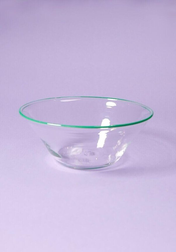 A clear glass bowl with a green rim on a lavender background.