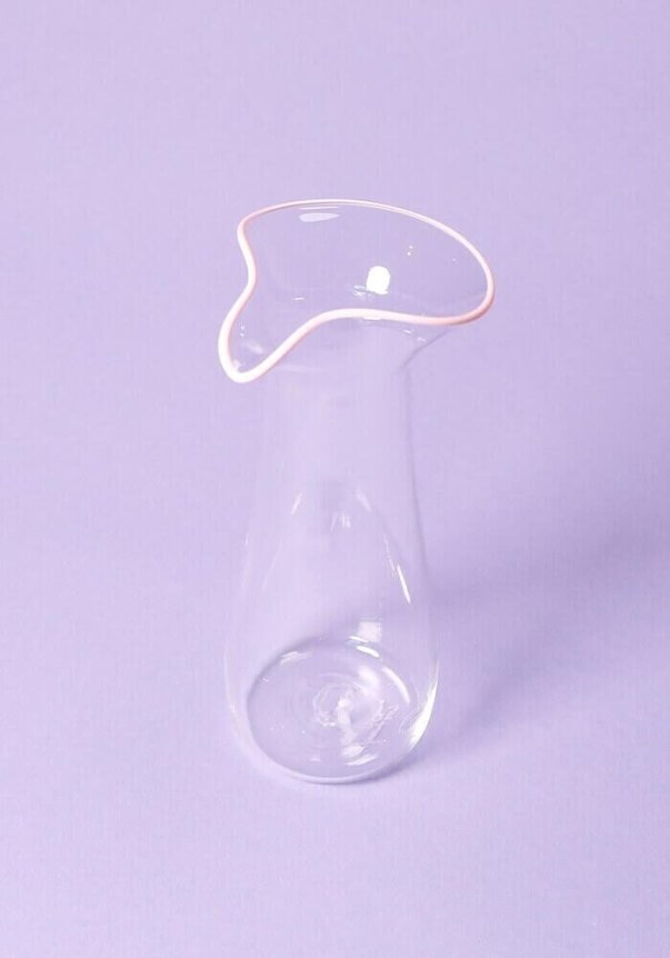 A clear glass carafe with a unique, asymmetrical rim design, set against a light purple background.