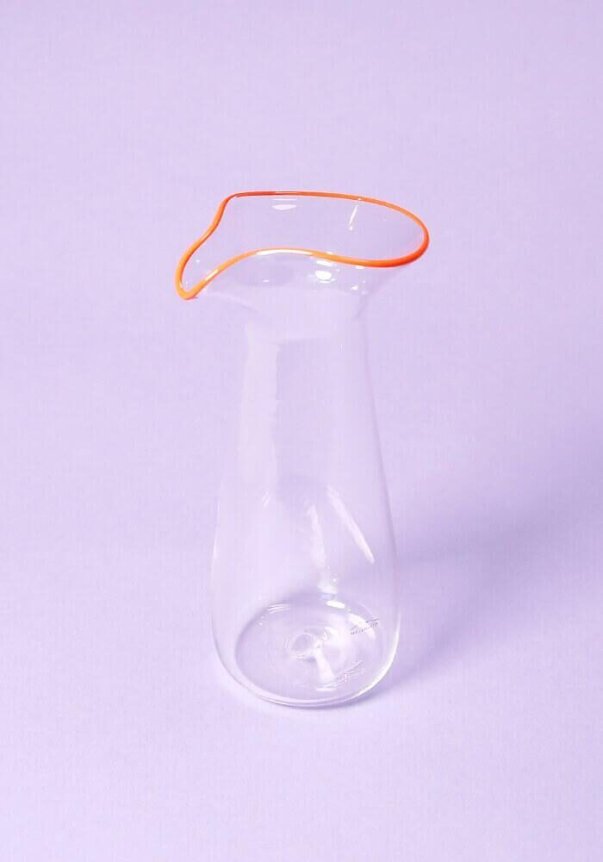 A clear glass carafe with an orange-rimmed wavy edge on a light purple background.