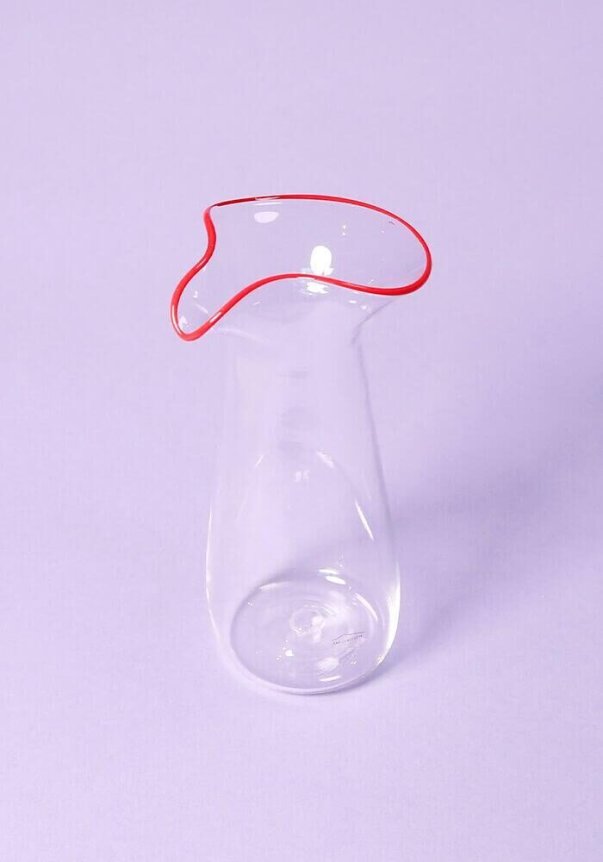 A clear glass vase with a uniquely shaped red-trimmed rim set against a light purple background.