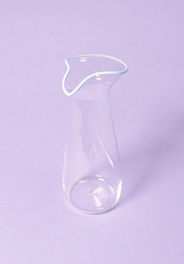 A clear glass carafe with a wavy spout, set against a pastel purple background.