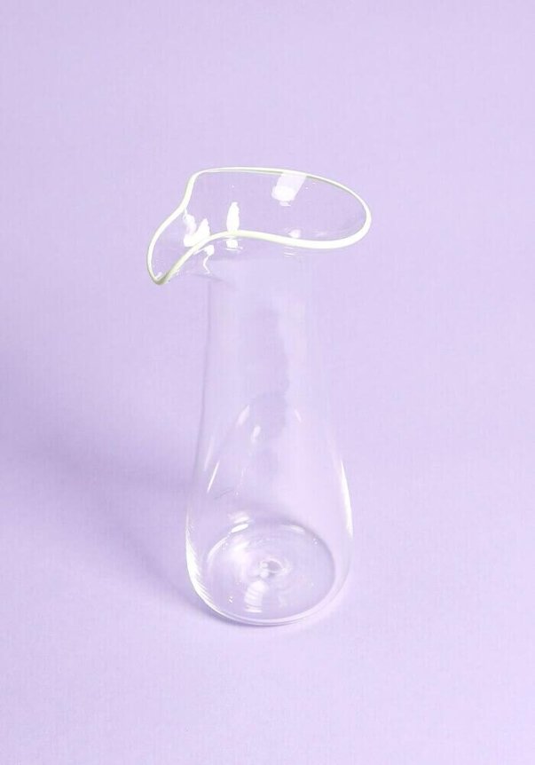 A clear glass pitcher with a uniquely curved spout on a light purple background.