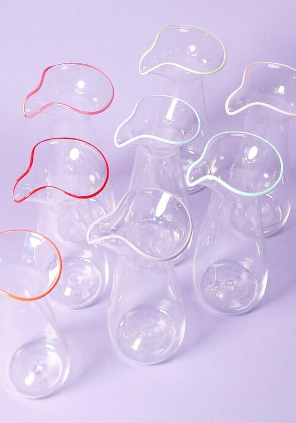 A collection of clear glass pitchers with colorful, wavy-edged rims set against a light purple background.