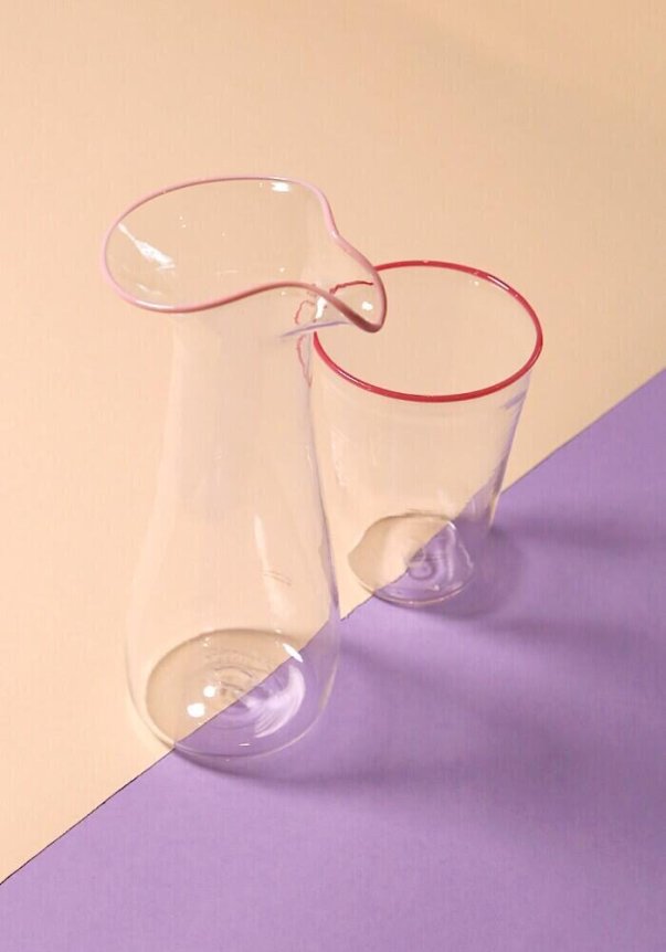 A clear glass pitcher and cup with pink rims are placed on a pastel beige and purple geometric background.