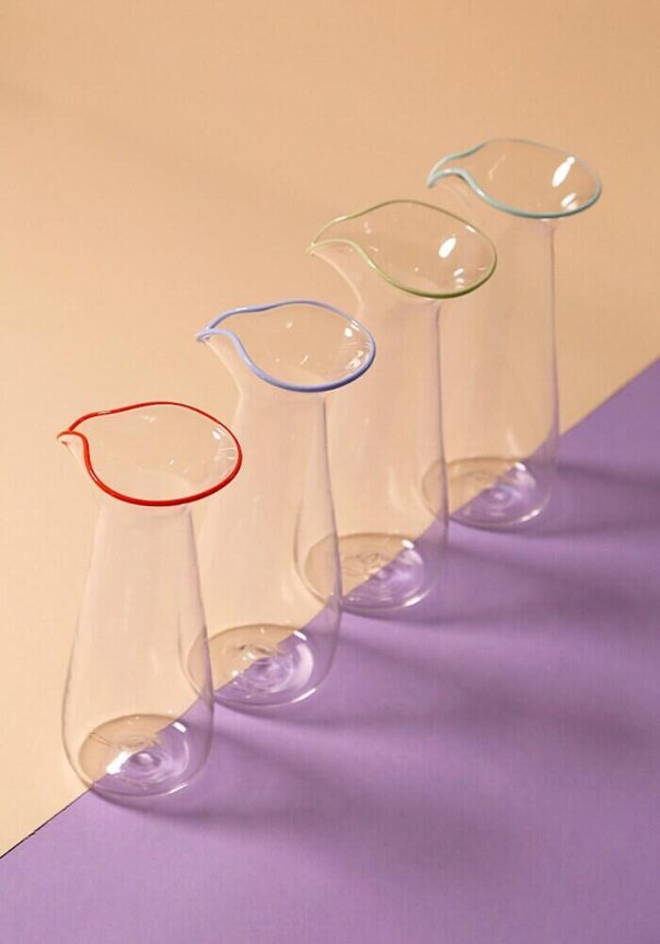 A set of four elegant glass carafes with colorful rims (red, blue, green, and teal) on a dual-toned beige and purple background.