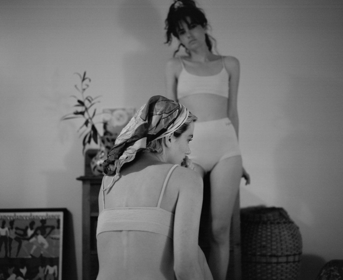 Two women dressed in simple, fitted, light-colored underwear, with one wearing a patterned headscarf, set against a minimalist background with floral decor.
