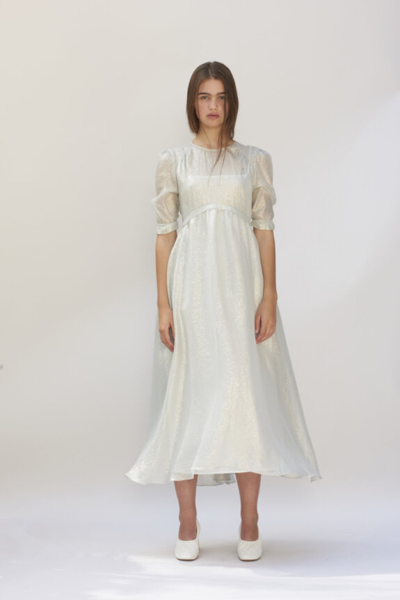 A model wearing a shiny, light-colored long dress with puff sleeves and white shoes, standing against a plain background.