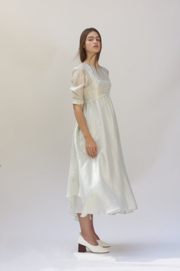 A woman is wearing a shimmery, light-colored midi dress with puff sleeves and white shoes, posing against a plain background.