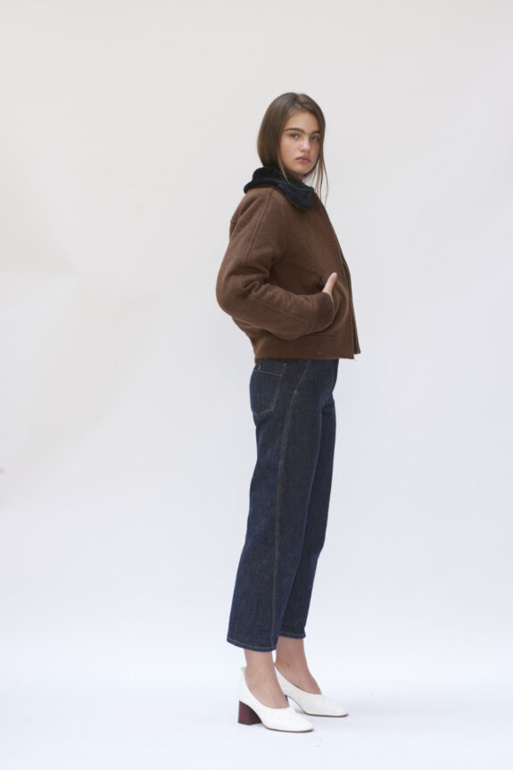 A woman stands in a brown jacket, dark jeans, and white heels against a plain background.