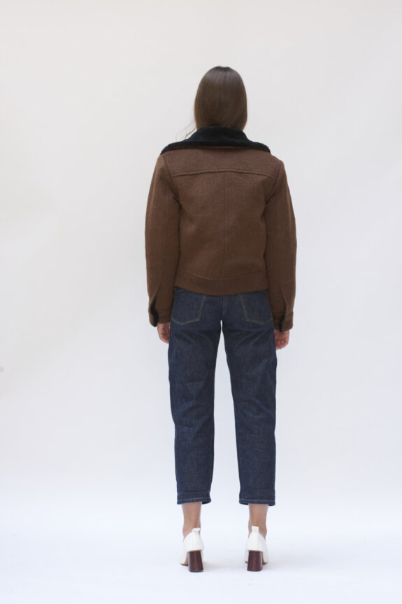 A person wearing a brown jacket and blue jeans stands facing away, with white heels visible.