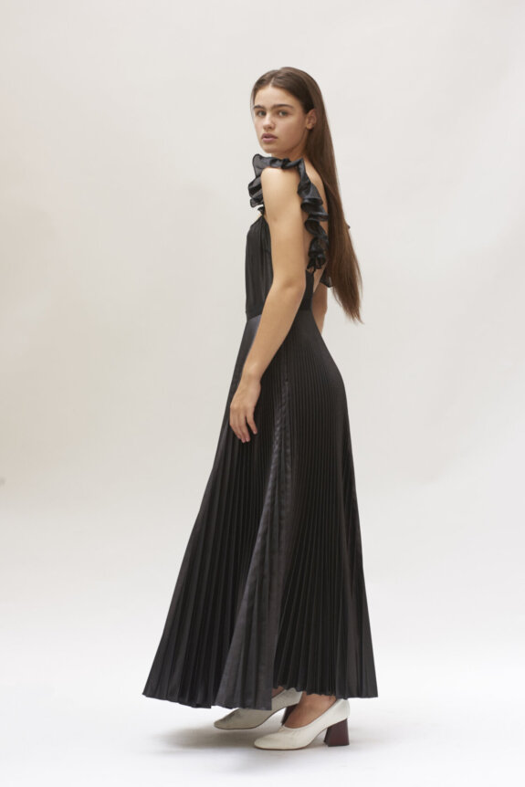 A woman wearing a long, pleated black dress with ruffled shoulders and white shoes stands, looking to the side.