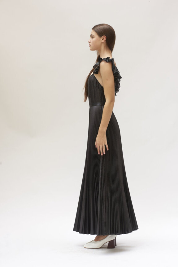 A woman in a black pleated dress and white heels stands in profile against a plain background.