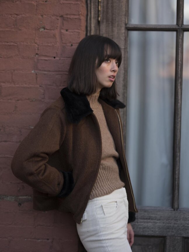 A person with shoulder-length hair leans against a brick wall, wearing a brown jacket over a beige sweater and white pants.