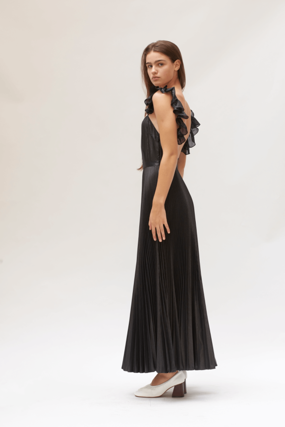A model wears a black pleated gown with ruffled straps and a fitted silhouette, paired with white block-heeled shoes.