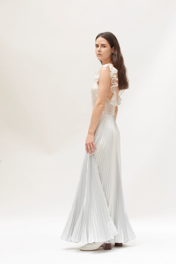 A model wearing a floor-length, silver, pleated gown with ruffled shoulder detailing, offering an elegant and flowing silhouette.