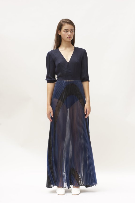 A model wearing a navy blue, sheer pleated maxi dress with a deep V-neck and three-quarter sleeves, paired with white shoes.