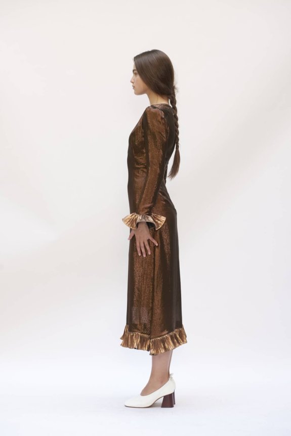 A side profile of a model wearing a fitted, shimmer brown long-sleeve dress with gold ruffle cuffs and hem, paired with white heeled shoes.