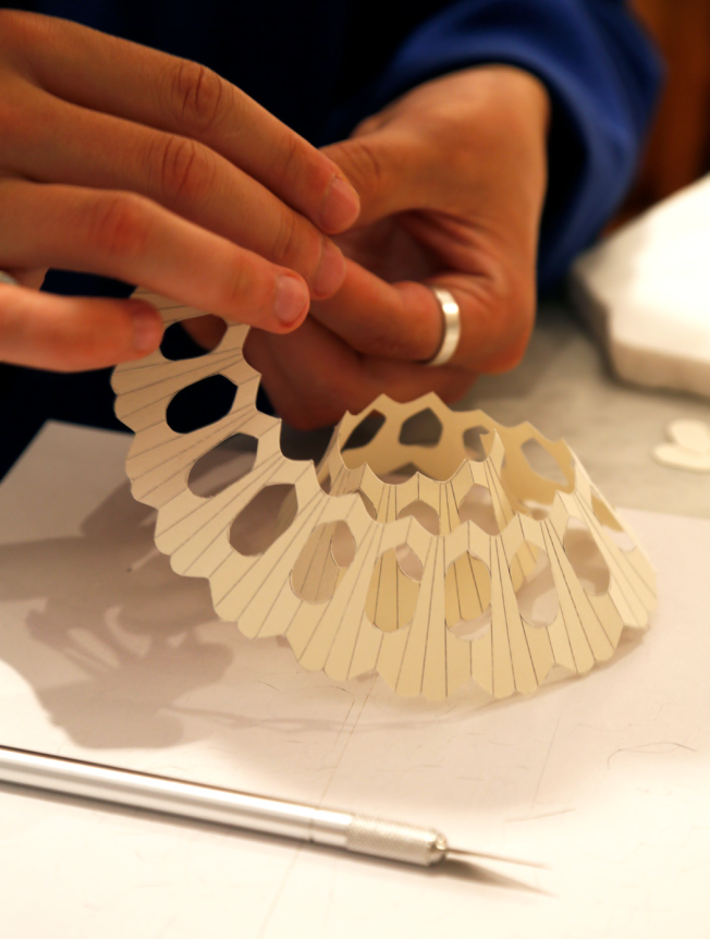 A person is crafting an intricate, geometric paper sculpture with a lattice design, using a precision knife on a white surface.