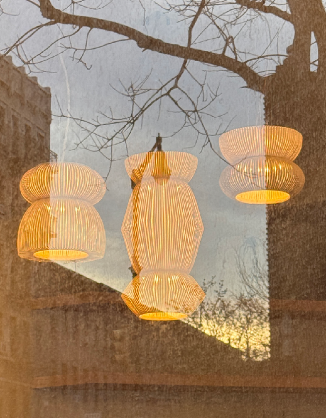 A trio of warmly lit, ribbed glass pendant lights reflecting against a window with tree branches and a sunset backdrop.