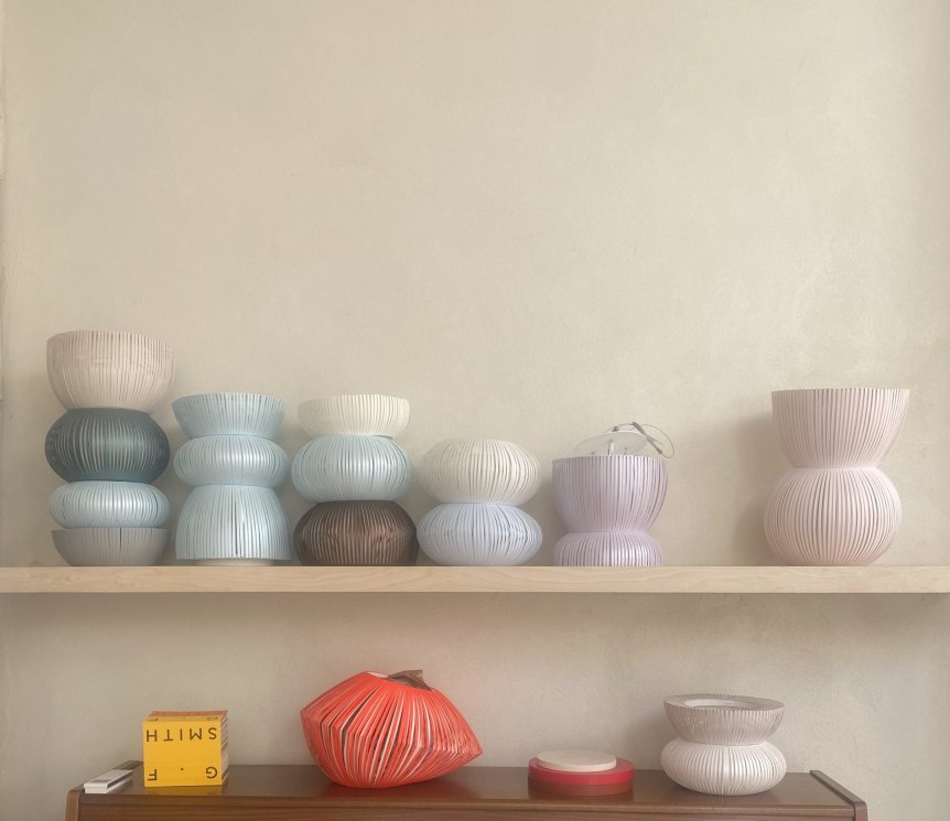 A collection of vertically-ribbed, stacked vases in soft pastel and neutral tones, displayed on wooden shelves against a light beige wall, alongside a bright orange ribbed object and a yellow cube with black lettering.