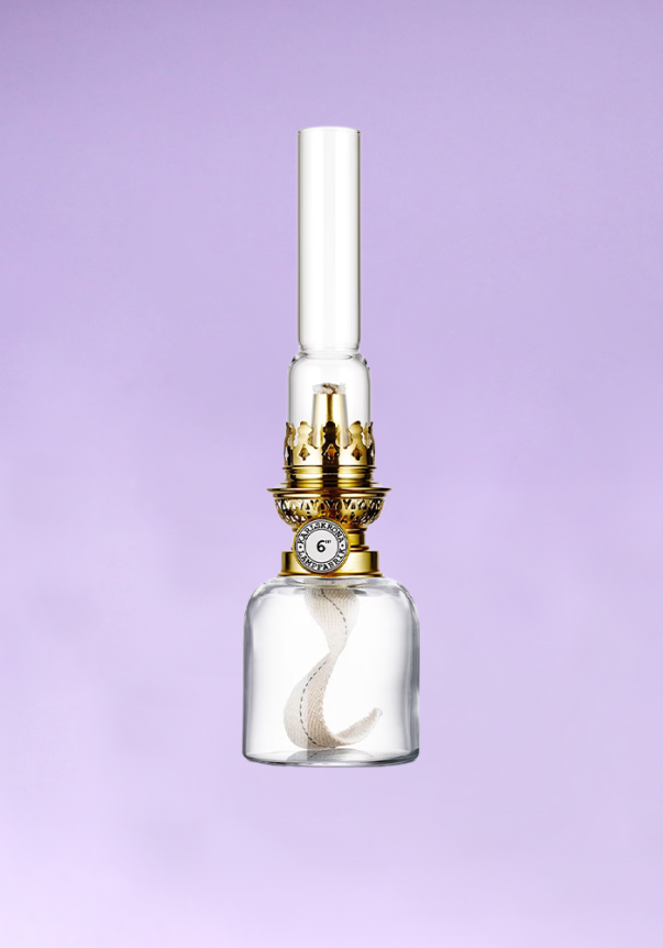 A vintage-style oil lamp with a glass body, gold accents, and a twisted fabric wick against a lavender background.