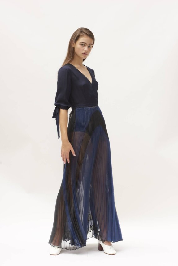 A woman is wearing a navy blue top with mid-length sleeves and a pleated, semi-transparent, long blue skirt. She is standing in a minimalist setting with white shoes.