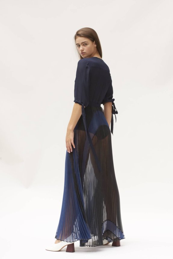 A woman is modeling a navy blue outfit with a sheer, pleated skirt that reveals dark shorts underneath. She is wearing white heels with brown block heels, and the background is a plain white.