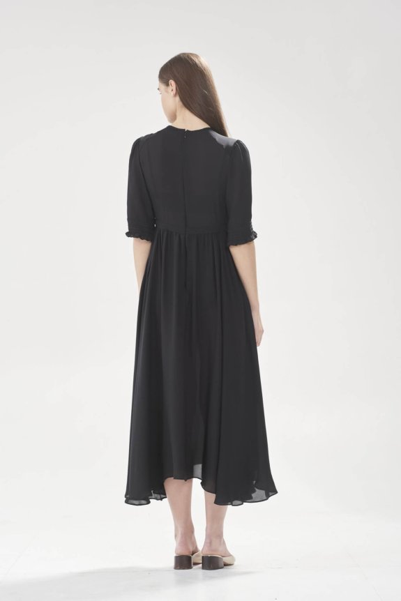 A woman with long brown hair is wearing a black, knee-length dress with half sleeves. She is facing away, standing against a plain white background.