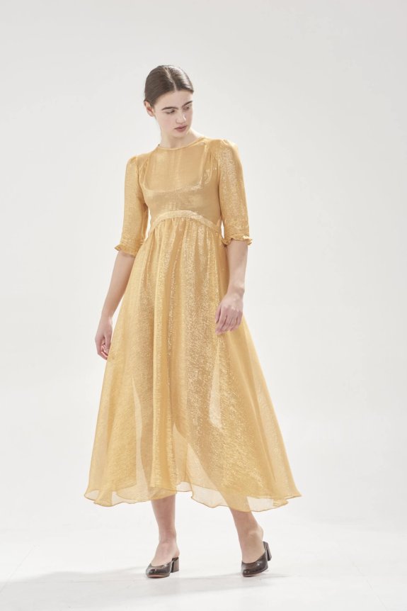A woman is wearing a shimmering gold dress with elbow-length sleeves, standing against a plain background. She has dark hair tied back and is looking downwards.