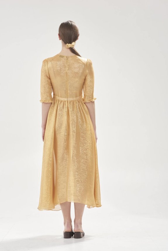 A woman is wearing a shimmering gold midi dress with short sleeves and standing with her back to the camera in a minimalist white space. Her hair is tied back with a matching scrunchie, and she is wearing black shoes.
