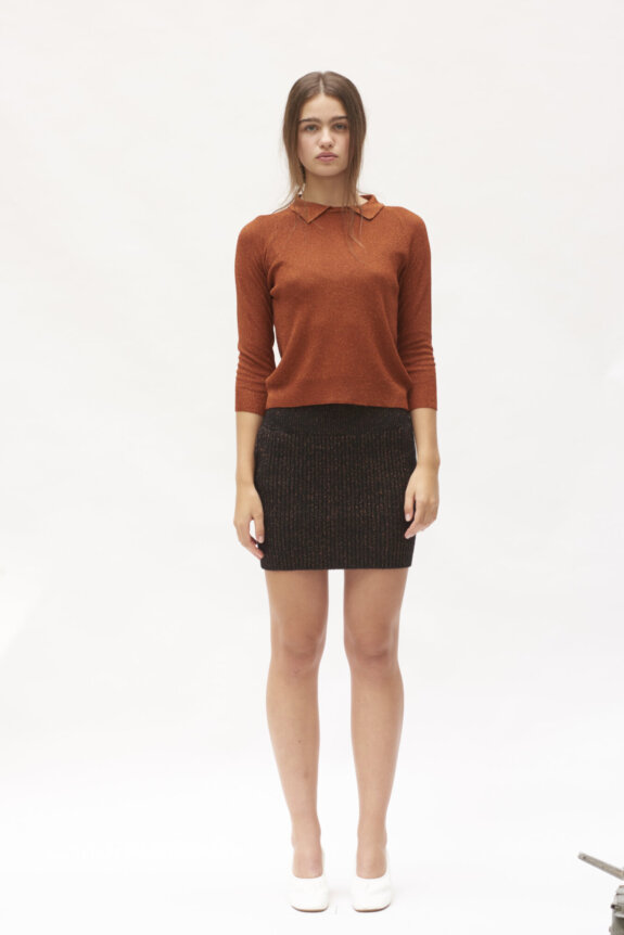 A woman wearing a rust-colored sweater, a dark skirt, and white shoes standing against a plain background.