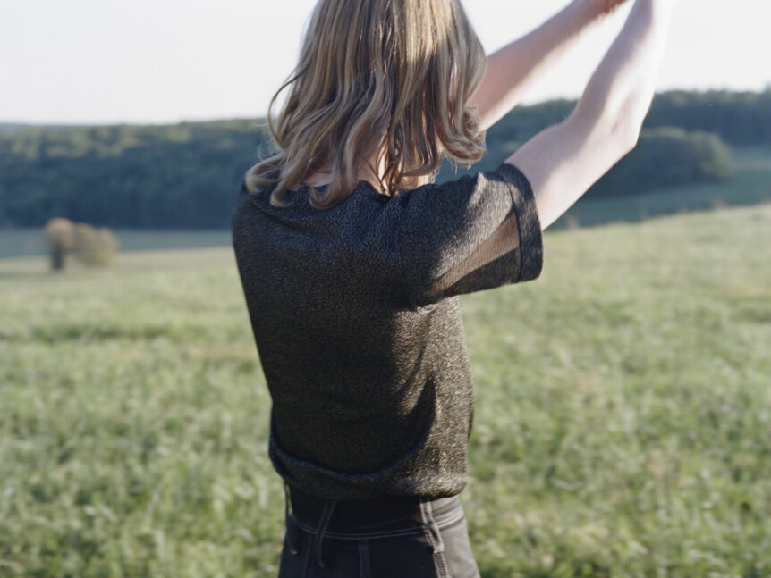 A person with shoulder-length hair is standing in a grassy field, facing away, with arms raised.
