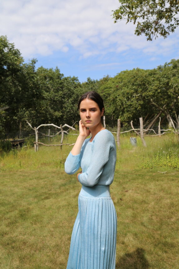 A woman in a light blue outfit standing on a grassy area with trees in the background.