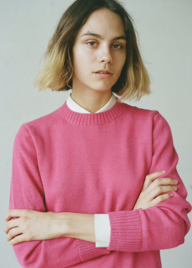 A person in a pink sweater poses with crossed arms and a neutral expression.
