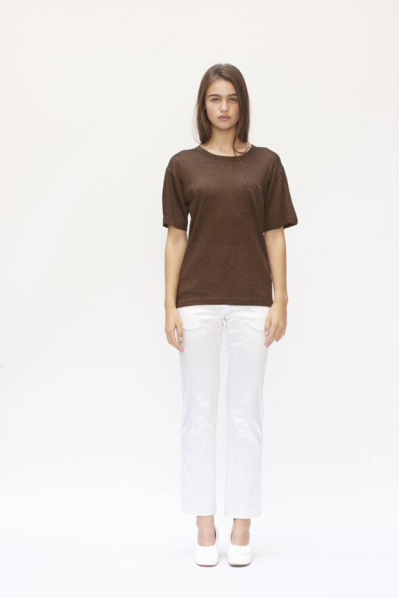 A woman wearing a brown top and white pants standing against a plain background.