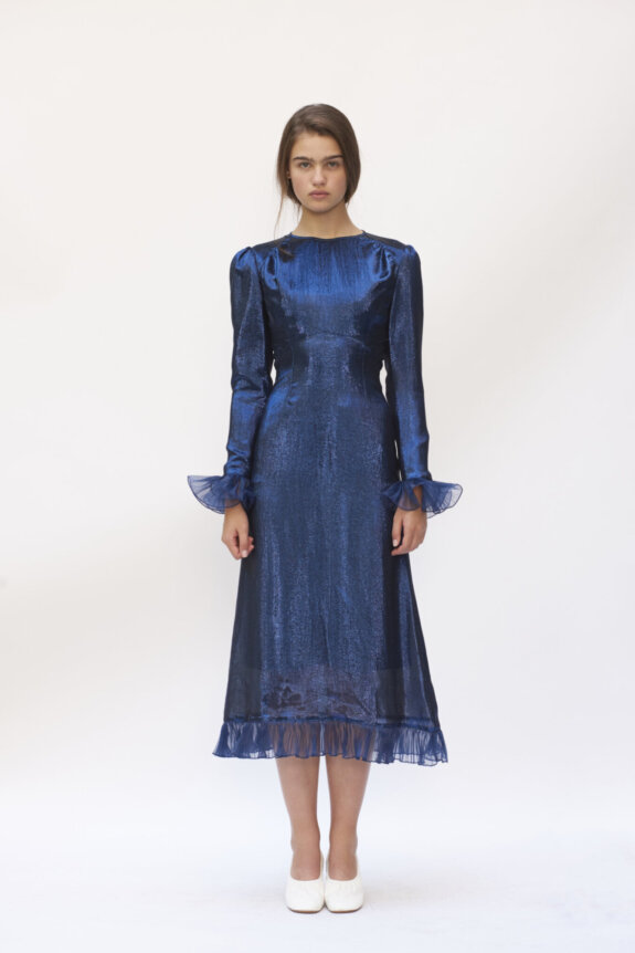 A model wearing a shimmering blue dress with ruffled cuffs and hem, paired with white shoes.