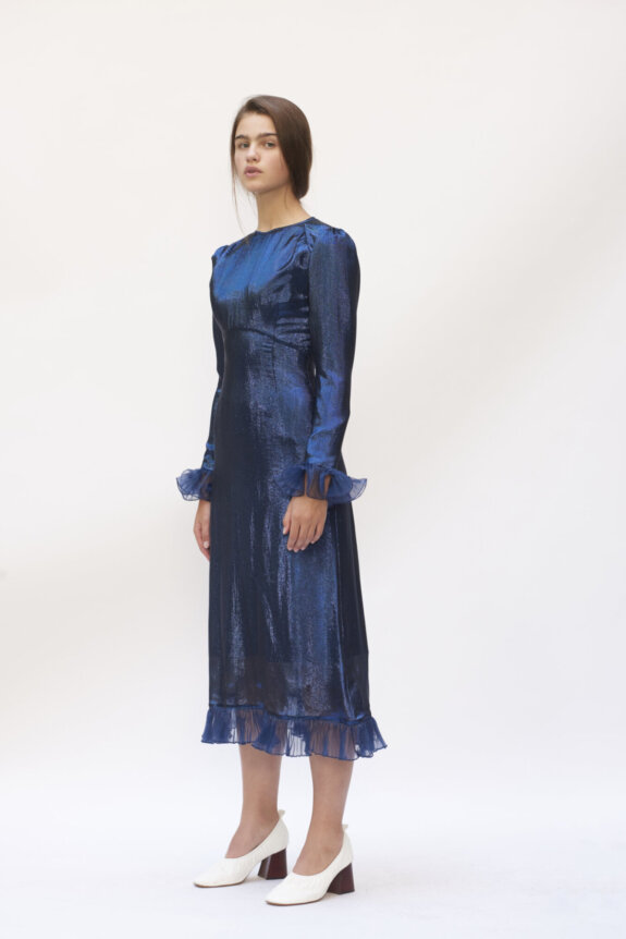 Woman wearing a shimmering blue dress with frilled cuffs and hem, paired with white block-heeled shoes.