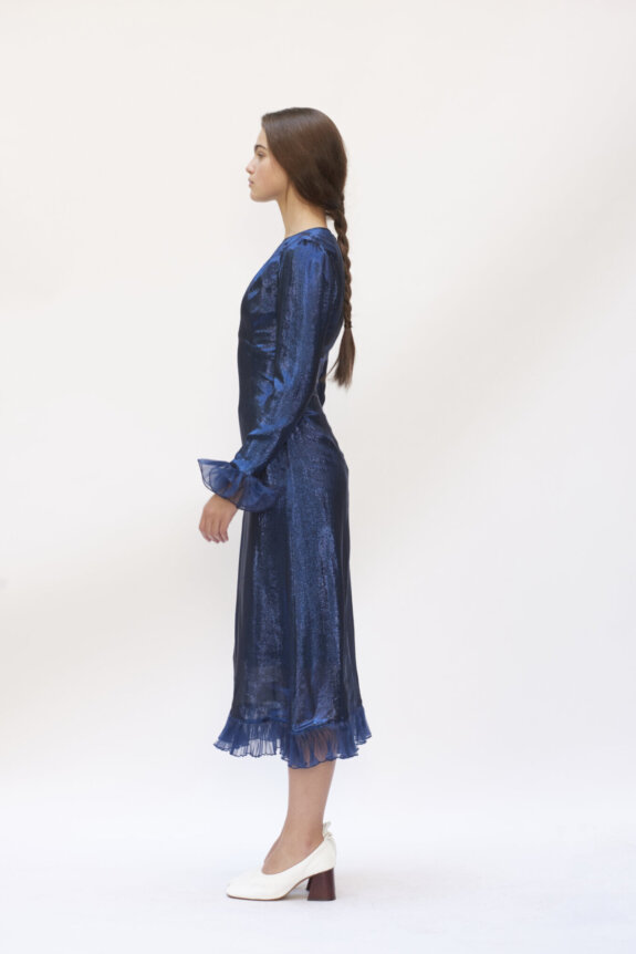 A woman in a shiny blue dress with long sleeves and a braid stands in profile, wearing white heeled shoes.