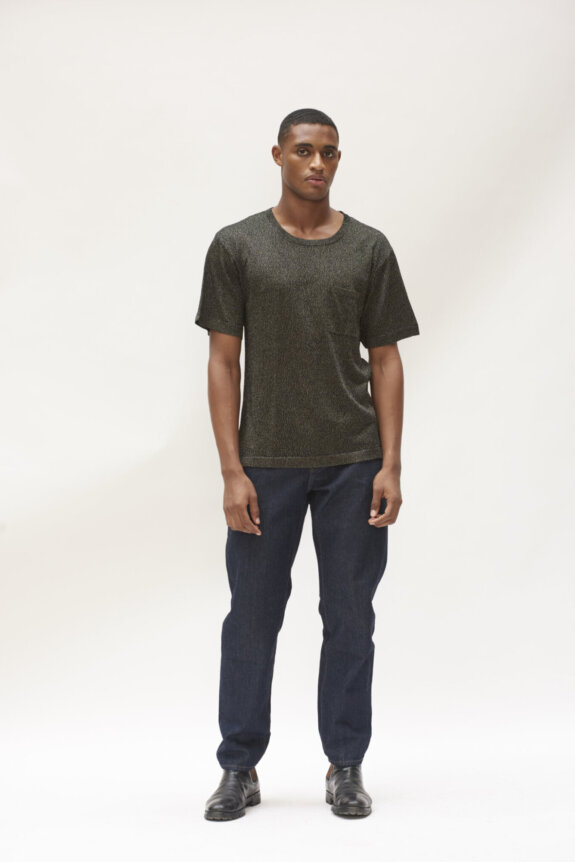 A man wearing a dark textured T-shirt, dark jeans, and black shoes standing against a light background.