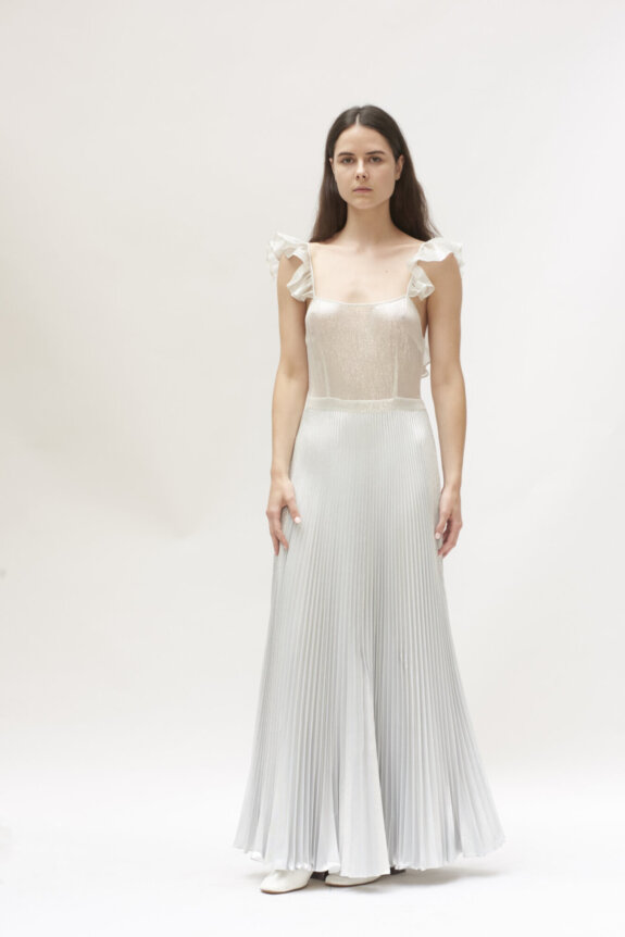 A woman in a light, flowing pleated dress with ruffle straps stands against a plain background.