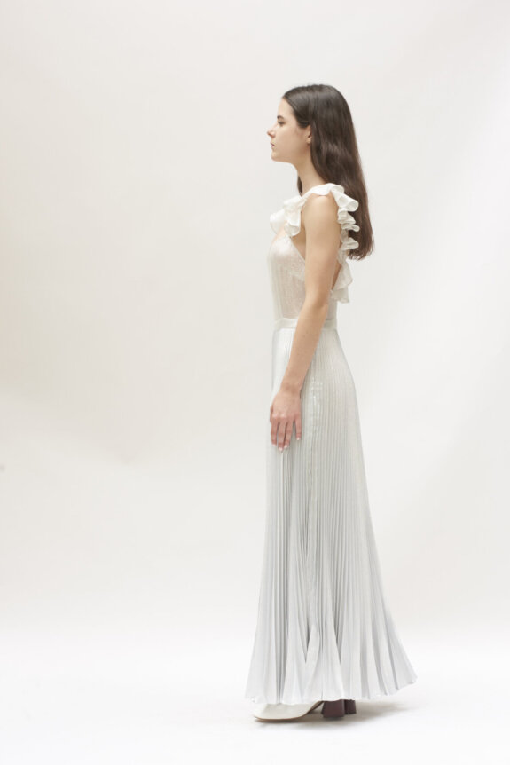 A woman in a white pleated dress with ruffled straps stands in profile against a plain background.