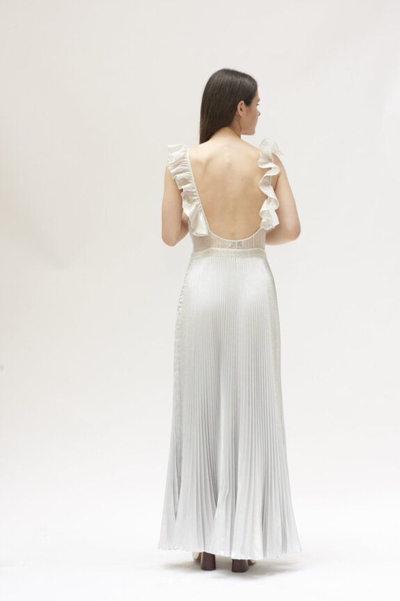 A woman wearing a backless, ruffled, and pleated silver dress stands facing away from the camera.