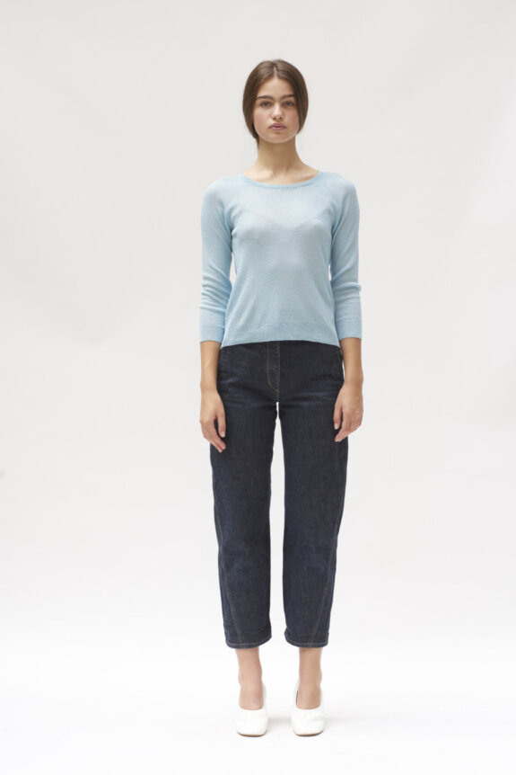 A person wearing a light blue top, dark jeans, and white shoes stands against a plain background.