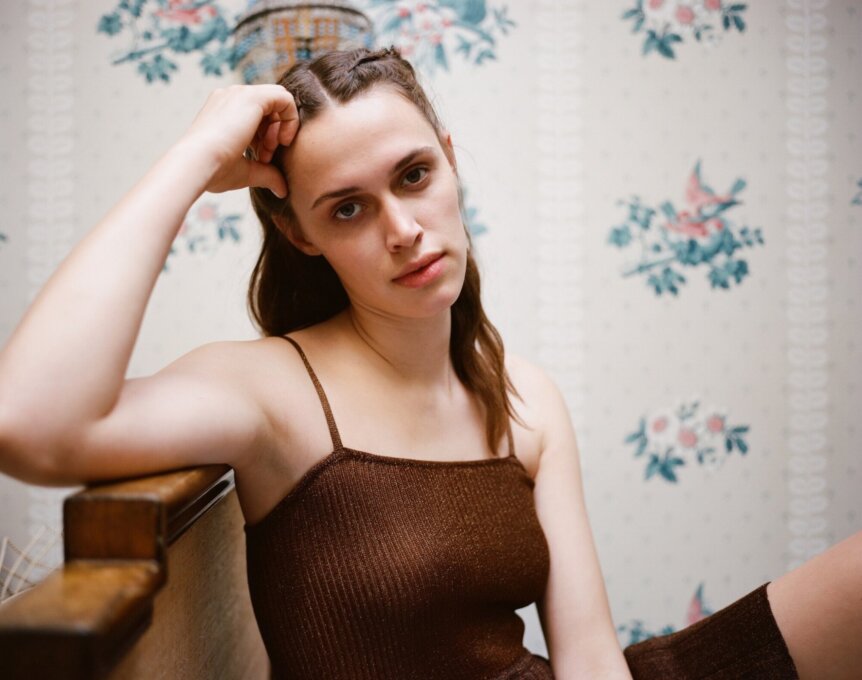 A person with long hair and a brown tank top sits on a bench against a floral wallpapered wall.