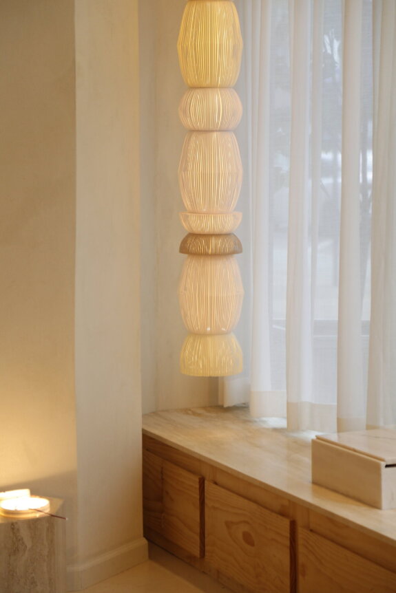 A stack of illuminated, uniquely shaped pendant lamps beside a window with sheer curtains.