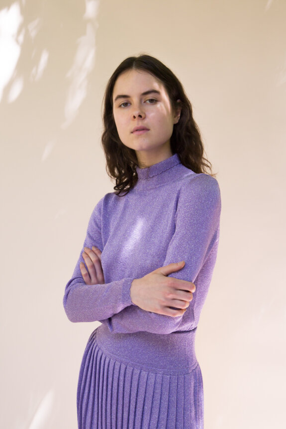 A woman in a lavender sparkly outfit stands with arms crossed against a light background.